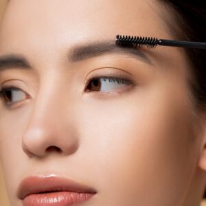Close up view of young asian woman with natural makeup applying eyebrow gel isolated on beige