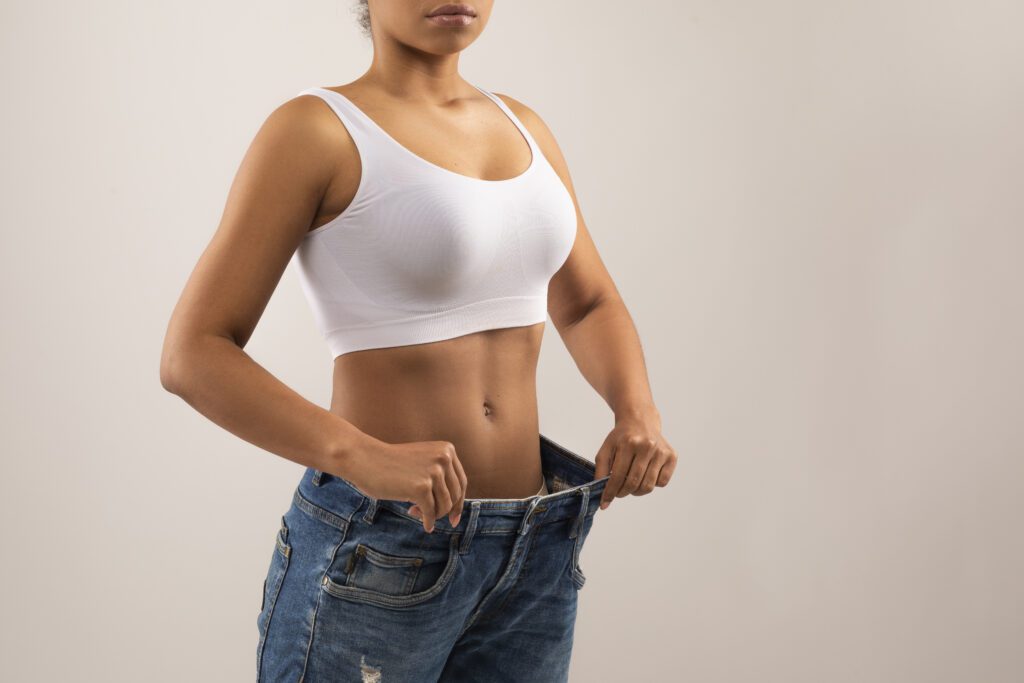 Diet, healthy lifestyle, slimming concept. Cropped of slim well-fit young black woman wearing white top and big jeans after weight loss diet program on grey studio background, copy space