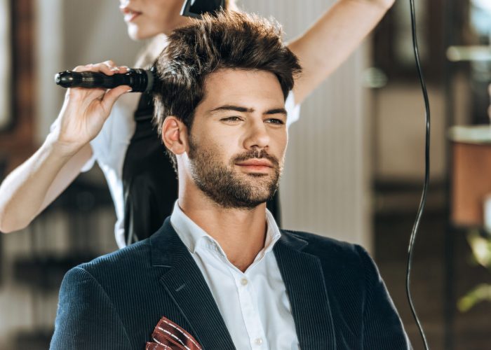 cropped shot of hairstylist combing and drying hair to handsome young man in beauty salon